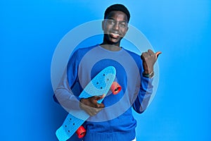 Young african american man holding skate pointing thumb up to the side smiling happy with open mouth