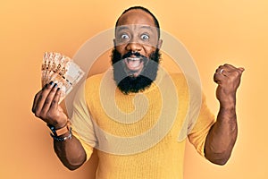 Young african american man holding south african 20 rand banknotes pointing thumb up to the side smiling happy with open mouth