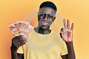 Young african american man holding south african rand banknotes doing ok sign with fingers, smiling friendly gesturing excellent