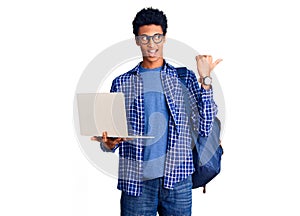 Young african american man holding student backpack using laptop pointing thumb up to the side smiling happy with open mouth