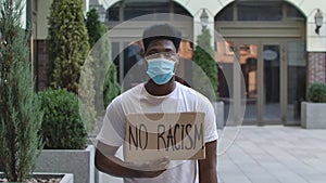 Young African American man in a medical mask stands with a cardboard poster NO RACISM in a public outdoor place. An anti