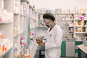 Young African American man pharmacist standing in interior of pharmacy. Man specialist of pharmacy making notes on