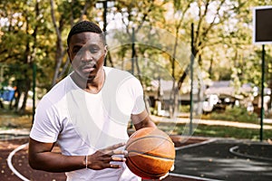 A young African American man is playing basketball on an outdoor court, smiling and looking at the camera. Outdoor