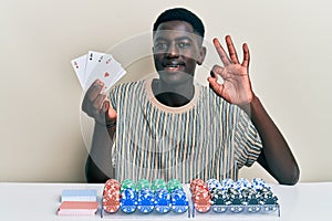Young african american man playing poker holding cards doing ok sign with fingers, smiling friendly gesturing excellent symbol