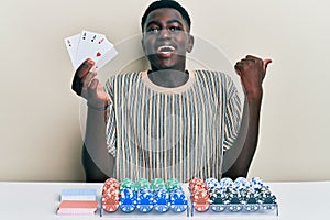 Young african american man playing poker holding cards pointing thumb up to the side smiling happy with open mouth