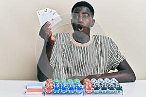 Young african american man playing poker holding cards scared and amazed with open mouth for surprise, disbelief face