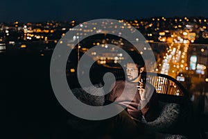 Young African AMerican man sitting on balcony with urban view and using tablet at night