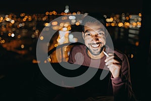 Young African AMerican man sitting on balcony with urban view and using tablet at night