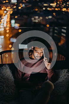 Young African AMerican man sitting on balcony with urban view and using tablet at night