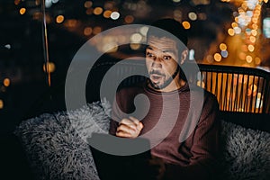 Young African AMerican man sitting on balcony with urban view and using tablet at night