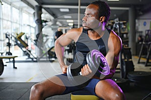 Young African American man sitting and lifting a dumbbell close to the rack at gym