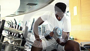 Young African American man sitting and lifting a dumbbell close to the rack at gym. Male weight training person doing a