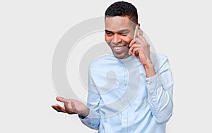 Young African American man talking on cellphone to his friend, looking cheerful and happy, posing on white studio background.