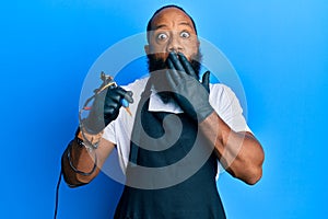 Young african american man tattoo artist wearing professional uniform and gloves holding tattooer machine covering mouth with