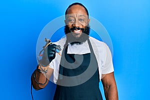 Young african american man tattoo artist wearing professional uniform and gloves holding tattooer machine looking positive and