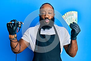 Young african american man tattoo artist wearing professional uniform and gloves holding tattooer machine and russian ruble