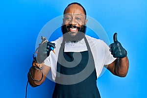 Young african american man tattoo artist wearing professional uniform and gloves holding tattooer machine smiling happy and