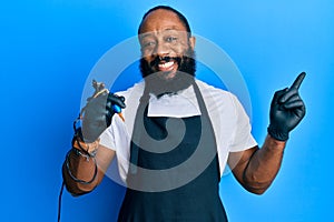 Young african american man tattoo artist wearing professional uniform and gloves holding tattooer machine smiling happy pointing