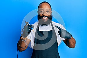 Young african american man tattoo artist wearing professional uniform and gloves holding tattooer machine smiling happy pointing