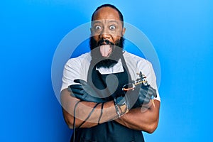 Young african american man tattoo artist wearing professional uniform and gloves holding tattooer machine sticking tongue out