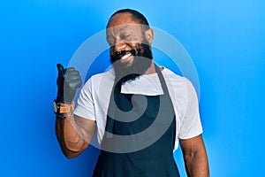 Young african american man tattoo artist wearing professional uniform and gloves pointing thumb up to the side smiling happy with