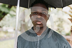 Young African American man under black umbrella in rain, sad. Fall or spring weather