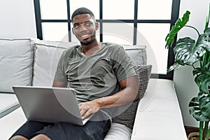 Young african american man using laptop at home sitting on the sofa looking away to side with smile on face, natural expression