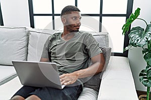 Young african american man using laptop at home sitting on the sofa looking to side, relax profile pose with natural face and