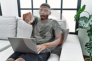 Young african american man using laptop at home sitting on the sofa pointing with finger surprised ahead, open mouth amazed