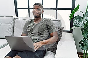 Young african american man using laptop at home sitting on the sofa smiling looking to the side and staring away thinking