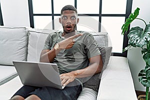 Young african american man using laptop at home sitting on the sofa surprised pointing with finger to the side, open mouth amazed