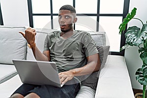 Young african american man using laptop at home sitting on the sofa surprised pointing with hand finger to the side, open mouth