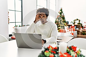 Young african american man using laptop sitting on the table by christmas tree doing ok gesture shocked with surprised face, eye