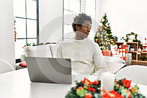 Young african american man using laptop sitting on the table by christmas tree looking to side, relax profile pose with natural