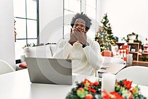 Young african american man using laptop sitting on the table by christmas tree shocked covering mouth with hands for mistake