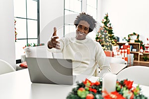 Young african american man using laptop sitting on the table by christmas tree smiling friendly offering handshake as greeting and