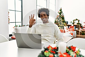 Young african american man using laptop sitting on the table by christmas tree smiling positive doing ok sign with hand and