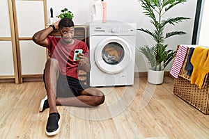 Young african american man using smartphone waiting for washing machine crazy and scared with hands on head, afraid and surprised