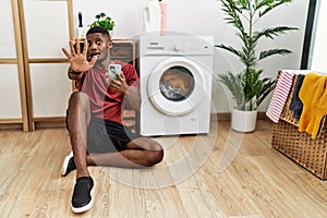 Young african american man using smartphone waiting for washing machine doing stop gesture with hands palms, angry and frustration