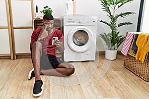 Young african american man using smartphone waiting for washing machine pointing to the eye watching you gesture, suspicious