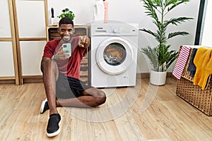 Young african american man using smartphone waiting for washing machine pointing to you and the camera with fingers, smiling