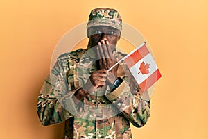 Young african american man wearing army uniform holding canadian flag covering mouth with hand, shocked and afraid for mistake