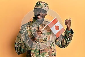 Young african american man wearing army uniform holding canadian flag pointing thumb up to the side smiling happy with open mouth