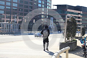 A young African American man wearing a black hoodie walking along a wide smooth sidewalk along a street with glass office building