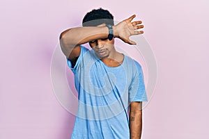 Young african american man wearing casual blue t shirt covering eyes with arm, looking serious and sad