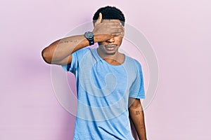 Young african american man wearing casual blue t shirt covering eyes with hand, looking serious and sad