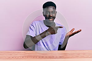 Young african american man wearing casual clothes sitting on the table amazed and smiling to the camera while presenting with hand