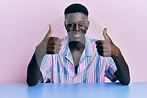 Young african american man wearing casual clothes sitting on the table approving doing positive gesture with hand, thumbs up