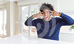 Young african american man wearing casual sweater sitting at home Doing peace symbol with fingers over face, smiling cheerful