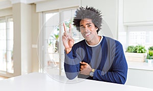Young african american man wearing casual sweater sitting at home smiling looking to the camera showing fingers doing victory sign